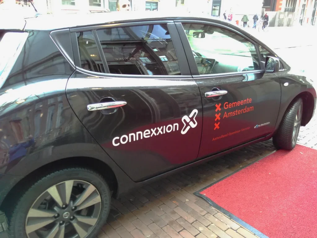 Elektrische Connexxion taxi in Amsterdam met blauwe kentekenplaat, taxiverzekering JW Verzekeringen 2026