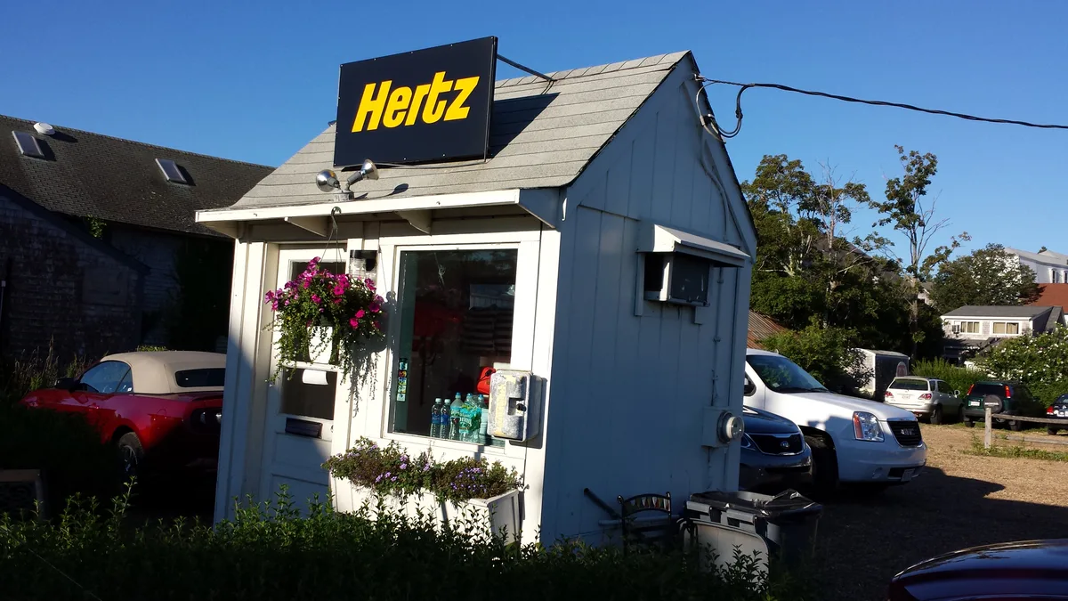 Huurauto verhuurkantoor met Hertz-bord en geparkeerde auto s — vergelijkbaar met aanbieders naast Sunnycars