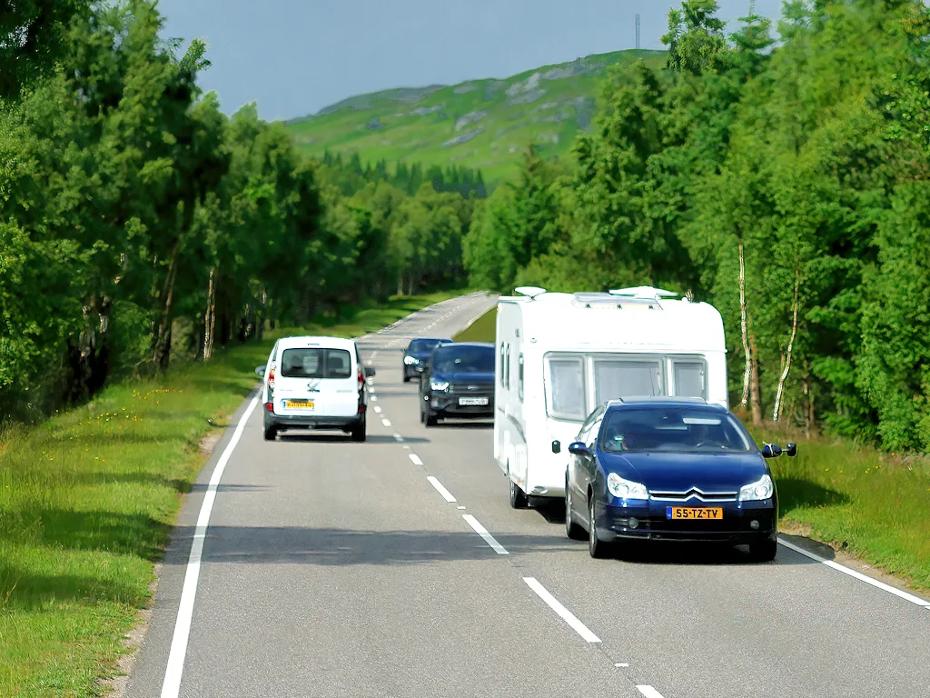 Auto met Nederlands kenteken trekt een witte caravan op een weg door groen landschap